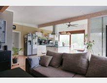 Offenes Wohnzimmer mit integrierter Küche in einem modernen Wohnhaus. Im Vordergrund steht ein graues Sofa, dahinter befinden sich eine helle Einbauküche, ein Esstisch mit Stühlen vor großen Fenstern und eine Holztrennwand mit sichtbaren Balken.