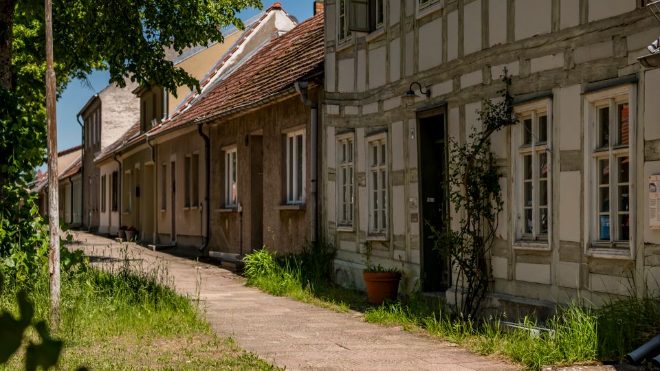 Schmale Wohnstraße mit Gehweg, gesäumt von niedrigen Häusern mit Satteldächern, teils als Fachwerkfassaden mit vielen Fenstern. Am Straßenrand stehen Gras und Bäume; die Gebäude liegen dicht aneinander und bilden eine geschlossene Häuserzeile.