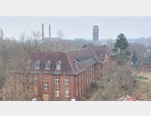 Backsteingebäude mit Satteldach in einer ländlichen Umgebung, umgeben von kahlen Bäumen. Im Hintergrund sind ein hoher Wasserturm sowie Industrieschornsteine sichtbar.