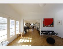 Wohnzimmer mit modernem Design, Holzboden in Fischgrätmuster, großen Fenstern und weißen Doppeltüren. Im Hintergrund ein Raum mit Bücherregalen, einem schwarzen Flügel und einem runden Tisch, vorne moderne Sessel und ein abstraktes Kunstwerk an der Wand.