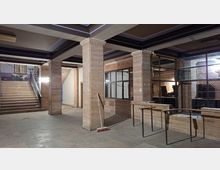 Innenraum einer verlassenen, industriell wirkenden Halle mit Betonboden, massiven Stützen und freiliegenden Deckenbalken. Links führt eine breite Treppe nach oben, rechts sind große Glasfenster und ein niedriger Tresenbereich mit Metallgeländer zu sehen.