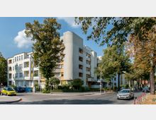 Mehrfamilienhäuser an einer Straßenkreuzung mit Bäumen im Vordergrund. Gebäude mit hellen Fassaden, gelben Fensterrahmen und Balkonen, umgeben von begrünten Vorgärten; Autos parken entlang der Straße.