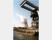 Ein Industriehafen mit einem Kühlturm eines Kraftwerks im Hintergrund, aus dem weißer Dampf austritt. Im Vordergrund liegt ein Frachtschiff an einem Kai neben einer großen Krananlage.