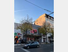 Straßenansicht eines modernen Theaters mit der Aufschrift "Astor Film Lounge" auf der Fassade. Vor dem Gebäude befinden sich Bäume, Laternen und parkende Autos, daneben sind ein Imbiss und ein Gebäude mit bemalter Fassade sichtbar.