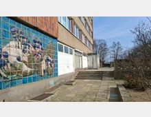 Außenansicht eines mehrstöckigen Wohn- oder Bürogebäudes mit verputzter Fassade und einem großen Wandmosaik aus blauen Fliesen. Davor liegt ein gepflasterter Gehweg mit Stufen und niedriger Betonbrüstung, umgeben von kahlen Bäumen und Sträuchern.