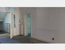 Ein leerer Innenraum mit weißen Wänden und einer geöffneten Glastür, die zu einem anderen Raum führt. Links befindet sich ein Heizkörper und eine Treppe mit einem weißen Geländer, während im Hintergrund ein weiterer Raum mit weißen Türen sichtbar ist.