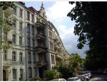 Straßenszene mit Gründerzeitfassaden, verzierten Balkonen und hohen Fenstern auf der linken Seite, umgeben von Bäumen; entlang der Straße sind geparkte Autos zu sehen, und der Himmel ist teils bewölkt.
