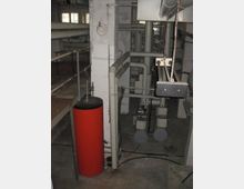 Kellerraum mit industrieller Heizungs- oder Rohrleitungsanlage. Sichtbar sind graue Rohre, Verbindungen, Ventile sowie ein roter, zylinderförmiger Behälter. Die Wände sind aus Beton, und der Boden ist schlicht und unbefestigt.