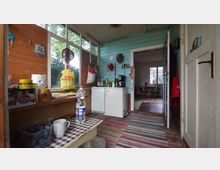 Eine kleine, rustikale Küche mit Holzwänden, einer Mischung aus hellblauer und naturholzfarbener Verkleidung, zahlreichen Regalen für Küchenutensilien und einem kleinen Kühlschrank. Große Fenster auf der linken Seite lassen Tageslicht herein, während der Boden mit bunten Teppichen bedeckt ist; im Hintergrund ist ein angrenzender Wohnraum sichtbar.