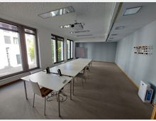 Heller Besprechungsraum mit langen weißen Tischen und Stühlen in der Mitte, großen Fenstern auf der linken Seite, grauen Wänden und einem an der Decke befestigten Projektor. An der rechten Wand hängt ein Regal mit bunten Kugeln, und im Raum befinden sich auch technische Geräte wie ein Luftreiniger.