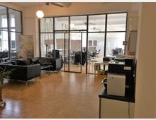 Offener Bürobereich mit modernen Glastüren, die einen Konferenz- oder Arbeitsraum mit mehreren Schreibtischen und Computern abtrennen. Im Vordergrund befinden sich schwarze Ledersessel, ein kleiner Tisch und ein Druckerbereich, alles auf einem Holzboden.