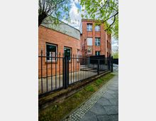 Backsteinfassade eines Gebäudes mit modernem, schwarzem Metallzaun im Vordergrund. Umgeben von Bäumen und Gehweg aus Kopfsteinpflaster.