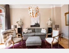 Wohnzimmer mit einem grauen Sofa und passendem Hocker auf einem dunkelroten Teppich, flankiert von zwei klassischen Sesseln. Zwei Marmorbüsten stehen auf schwarzen Sockeln neben dem Sofa, darüber hängt eine moderne, skulpturale Deckenleuchte, mit einem gerahmten Schwarz-Weiß-Bild an der Wand im Hintergrund.