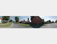 Panoramafoto einer ländlichen Straßenszene mit einer roten Backsteinkirche und Fachwerkturm auf der rechten Seite. Links stehen Wohnhäuser mit roten Dächern und grünen Vorgärten, während Bäume und ein breiter Gehweg die Straße säumen.