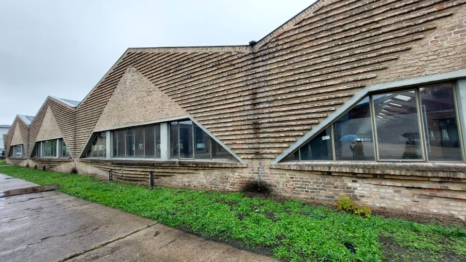 Backstein-Gebäude mit schräg verlaufenden Ziegelreihen und großen, dreieckigen Fenstern, die in regelmäßigen Abständen angeordnet sind. Im Vordergrund eine Asphaltfläche und Grünflächen, im Hintergrund bewölkter Himmel.