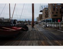 Holzpier mit festgemachten Segelbooten neben modernen Backsteingebäuden; im Hintergrund die Oberbaumbrücke und Stadtarchitektur bei bewölktem Himmel.