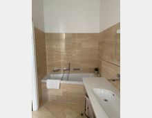 Ein Badezimmer mit einer Badewanne und einem Waschbecken, umgeben von beigefarbenen Fliesen. Links der Badewanne hängt ein weißes Handtuch, daneben befindet sich eine kleine Pflanze auf der Ablage über dem Waschbecken.