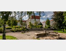 Ein kleiner Park mit Sandspielplatz und moderner Rutsche, umgeben von viel Grün, Bänken und einer Treppe, die zu höher gelegenen Bereichen führt. Im Hintergrund sind Wohngebäude mit Balkonen sichtbar.
