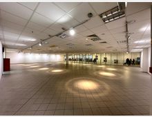 Leerer Innenraum eines Gebäudes mit gefliesten Böden, einer Rasterdecke mit eingebauten Leuchten und weißen Wänden. Im Hintergrund befinden sich große Glasfenster mit Blick auf einen belebten Bereich mit Menschen.