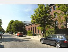 Straße mit Kopfsteinpflaster, gesäumt von Bäumen und geparkten Autos, rechts ein langes Backsteingebäude mit zahlreichen Fenstern und begrünten Fassaden. Im Hintergrund ist ein Teil eines Daches mit großen Glasflächen sichtbar.