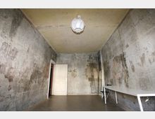 Ein minimalistischer Raum mit rauen, unverputzten Betonwänden und einer einfachen Deckenlampe. Der Raum enthält einen weißen Tisch, einen Stuhl und einen angelehnten Spiegel; eine Tür führt in einen angrenzenden Bereich.