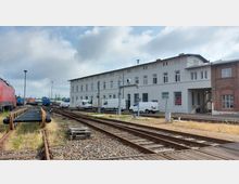 Bahnhofsgelände mit Gleisen, einem roten und mehreren blauen Zugwaggons im Hintergrund. Auf der rechten Seite steht ein langes, hellgraues Gebäude mit mehreren Fenstern und geparkten weißen Lieferwagen, daneben ein älteres Backsteingebäude.