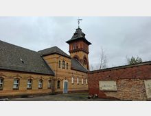 Backsteinbau mit langem, niedrigem Gebäudetrakt und einem hohen Turm mit dunkler Dachhaube und Wetterfahne. Im Vordergrund liegt ein asphaltierter Hof; rechts steht eine niedrige Mauer mit teils freigelegtem Mauerwerk unter grauem, bewölktem Himmel.