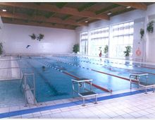 Hallenbad mit einem Schwimmbecken, das in Bahnen unterteilt ist, umgeben von gefliesten Böden. Große Fenster an der rechten Seite lassen Tageslicht herein, und einige Topfpflanzen und Wanddekorationen sorgen für eine freundliche Atmosphäre.