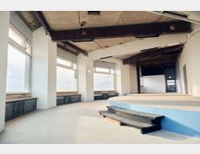 Industrieller Innenraum mit großen Fenstern, einem abgenutzten Betonboden und freiliegenden Deckenstrukturen. Im Vordergrund befindet sich eine runde Bühne mit schwarzen Stufen, im Hintergrund eine einfache Tür und eine Wand.