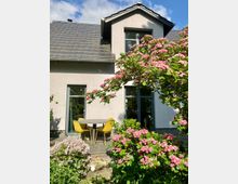 Ein modernes Einfamilienhaus mit hellgrauer Fassade und einem Schieferdach, umgeben von einem blühenden Garten mit rosa Blüten. Im Vordergrund steht eine Terrasse mit einem runden Tisch und gelben Stühlen.