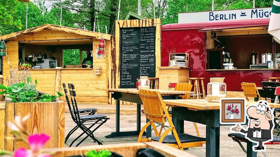 Ein Außenbereich eines kleinen gastronomischen Betriebs mit Holzmöbeln und einer Holzhütte, neben einem roten Verkaufswagen. Im Hintergrund befindet sich eine Speisetafel mit einer handgeschriebenen Menüauswahl. Umliegende Pflanzendekoration und grüne Bäume schaffen eine einladende Atmosphäre.