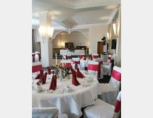 Festlich dekorierter Bankettsaal mit runden Tischen, weißen Tischdecken und roten Dekoelementen wie Servietten und Stuhlschleifen. Im Hintergrund ist ein erhöhter Bereich mit einer weißen Tafel und Blumenschmuck zu sehen, umgeben von klassischer Innenarchitektur mit verzierten Decken und Wandleuchten.