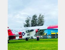 Ein kleines, weiß-rotes Propellerflugzeug mit der Aufschrift "NAVY" und der Nummer "862" steht auf einer Wiese. Im Hintergrund sind Bäume sowie ein blaues Gebäude mit gelben Markisen zu sehen.