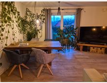 Wohnzimmer mit einem rustikalen Holztisch und vier Stühlen, umgeben von Pflanzen, die an der Wand hochranken. Im Hintergrund bodentiefe Fenster mit Blick auf einen Garten, daneben ein Fernseher auf einem niedrigen Holztisch mit Körben darunter.