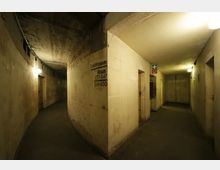Ein unterirdischer Betonbunker mit schmalen, gewölbten Gängen und kahlen Wänden. An einer Wand ist die Beschriftung "Sanitätsraum" mit Raumnummern sichtbar, Beleuchtung erfolgt durch einfache Deckenlampen.