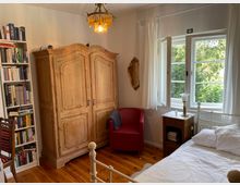 Ein gemütliches Schlafzimmer mit Holzboden, einem großen, rustikalen Holzschrank, einem Bücherregal, einem roten Sessel und einem weißen Metallbett. Neben einem Holz-Nachttisch mit Lampe befindet sich ein Fenster mit Blick auf grüne Bäume, umrahmt von weißen Vorhängen.
