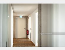 Ein heller, moderner Flur mit weißen Wänden, Holzboden und mehreren Türen. Am Ende des Flurs befindet sich eine geschlossene Tür mit einem leuchtenden Notausgangsschild darüber, daneben hängt ein Feuerlöscher an der Wand.