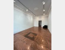 Ein leerer Raum mit glänzendem braunem Fliesenboden, einer weißen Decke mit Stuckverzierungen und eingebauten Strahlern. Im Hintergrund befindet sich ein schwarzer Polstersessel an der rechten Wand sowie eine Türöffnung, die zu einem anderen Raum führt.