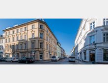 Straßenecke mit historischen Wohn- und Geschäftshäusern, eines in beigefarbener und eines in weißer Fassade, beide mit klassischen Stuckelementen. Im Vordergrund stehen geparkte Autos, die Straße ist gepflastert, und der Himmel ist klar und blau.