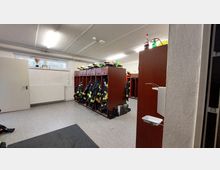 Umkleideraum mit Schränken aus Holz, in denen Feuerwehrschutzkleidung und Helme gelagert sind. Der Raum hat grauen Bodenbelag, eine weiße Wand mit Heizkörper und ein Fenster mit Deko-Elementen.
