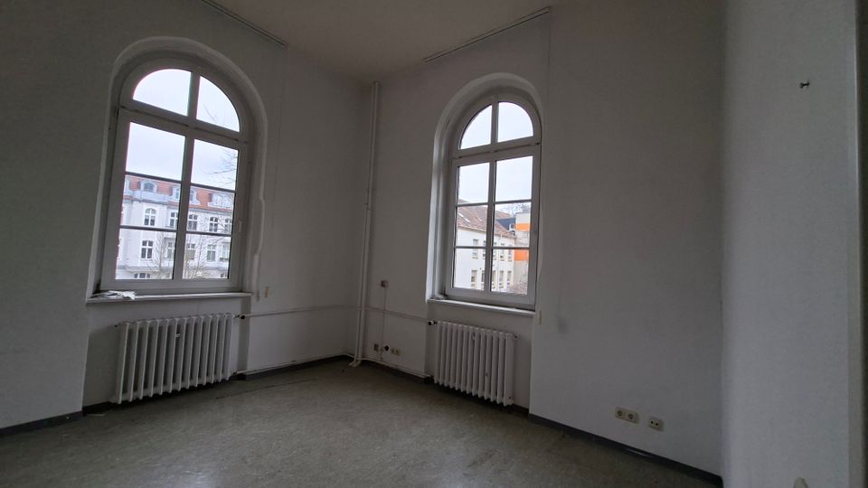 Leerer, heller Innenraum mit weißen Wänden und zwei großen, rundbogigen Sprossenfenstern, darunter jeweils ein Heizkörper. Grauer Boden und sichtbare Leitungen entlang der Wand; durch die Fenster sind gegenüberliegende Wohngebäude zu sehen.