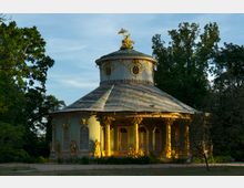 Pavillon im Rokokostil mit grünlicher Fassade, goldverzierten Säulen und Figuren, umgeben von Bäumen und einer Wiese. Auf dem Dach befindet sich eine goldene Figur mit Schirm.