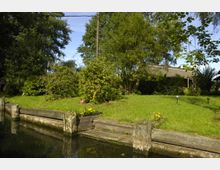 Ein grüner Garten mit vereinzelten Bäumen und Sträuchern, angrenzend an einen Wasserkanal mit hölzernen Uferbefestigungen. Im Hintergrund ist ein Haus mit Reetdach teilweise von Vegetation verdeckt.