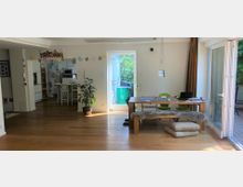 Wohnbereich in einem modernen Haus mit hellem Holzboden, einem Holztisch mit grünen Stühlen und einer Holzbank. Im Hintergrund eine offene Küche und eine Glastür, die zu einer Terrasse und einem Garten führt; Dekorationen und Alltagsgegenstände sind sichtbar.