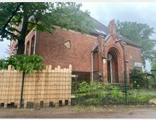 Historisches Backsteingebäude mit runden und dreieckigen Fenstern sowie einem spitz zulaufenden Eingangsportal. Vor dem Gebäude befinden sich ein Zaun, Holzbretter sowie Pflanzen und ein Baum.