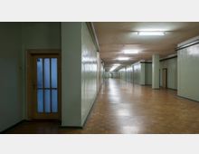 Langer, schlichter Flur mit grün gestrichenen Wänden, hölzernem Parkettboden und mehreren Türen auf beiden Seiten. Die Beleuchtung erfolgt durch an der Decke angebrachte Leuchtstofflampen.