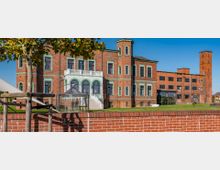 Backsteingebäude im historischen Stil mit grün umrahmten Fenstern und einem angebauten Turm, daneben ein modernerer Backsteinanbau mit großen Fenstern. Im Vordergrund eine niedrige Backsteinmauer, ein Glasdach, sowie ein Baum, umgeben von Rasenflächen unter klarem Himmel.