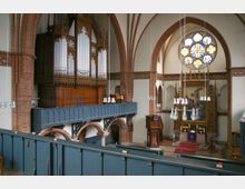 Innenraum einer Kirche mit gotischen Stilelementen. Zu sehen sind eine große Pfeifenorgel auf einer Empore, ein Altar mit einem Gemälde und aufwendig dekorierte Rundbogenfenster mit buntem Glas.