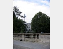 Steinerne Balustrade mit Blick auf den Berliner Fernsehturm, umgeben von Bäumen und modernen Gebäuden im Hintergrund, unter einem bewölkten Himmel.