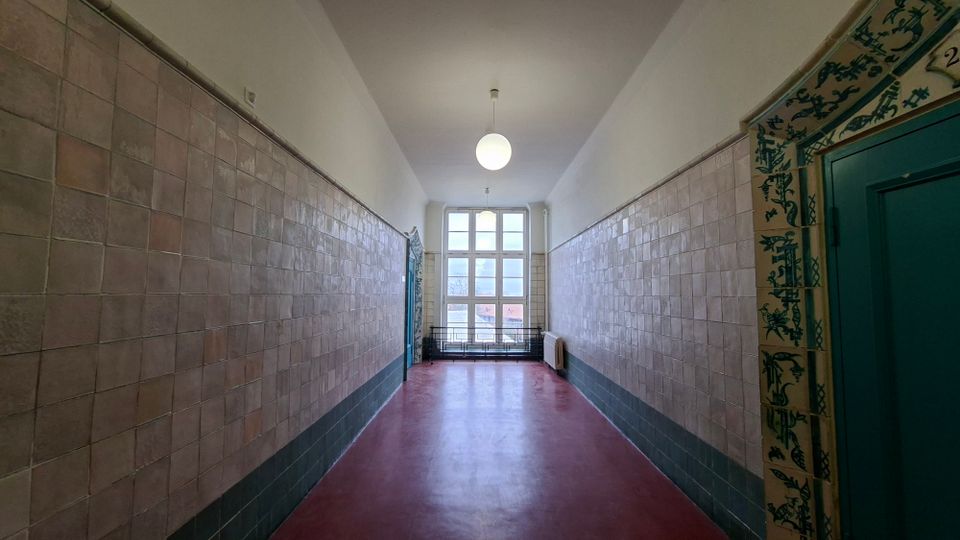 Langer, schmaler Innenflur mit rotbraunem Boden, hellen Fliesenwänden und hohen weißen Decken mit runden Hängelampen. Am Ende befindet sich ein großes, rechteckig unterteiltes Fenster; rechts ist eine türkisfarbene Tür mit dekorativem Fliesenrahmen zu sehen.